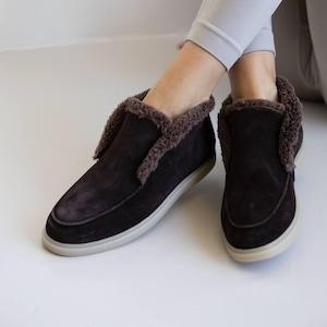 Op de afbeelding: Donkerbruine su&egrave;de instappers met een lichtgekleurde zool en een zachte, bruine imitatiebontvoering rond de opening. De schoenen zijn ontworpen voor comfort en warmte, geschikt voor binnen of casual wear.