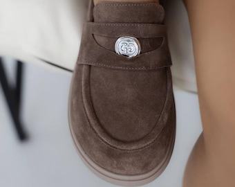 Könnte beinhalten: Braune Wildleder-Loafer mit silberfarbener Zierschnalle. Die Schuhe haben eine runde Spitze und eine flache Sohle. Das Design hat einen Riemen über dem Fuß mit einer kreisförmigen Schnalle. Die Loafer sind in einer neutralen Farbe gehalten.