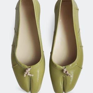 Op de afbeelding: Een paar olijfgroene ballerina's met een split-teenontwerp. De schoenen hebben een glanzende afwerking en een klein geknoopt detail bij de teen. De binnenkant is lichtbeige.