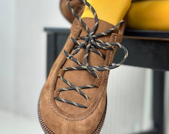 Puede incluir: Zapato de ante marrón con cordones negros y beige. El zapato tiene una puntera redondeada y una suela cosida. Los cordones están atados en forma de cruz. El zapato está sobre una superficie negra. La persona lleva medias amarillas.