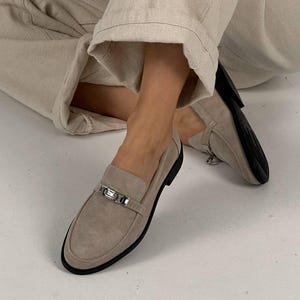 May include: Beige suede loafers with a silver buckle detail. The loafers have a pointed toe and a low heel.