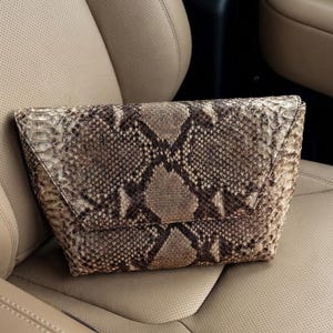 May include: A structured envelope-style clutch bag featuring a textured snakeskin pattern in shades of brown, tan, and cream. The bag has a pointed flap closure and a wide, tapered base, showcasing a detailed scale texture across the entire exterior.