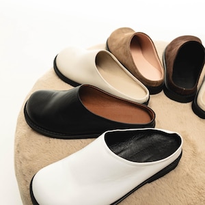 Op de afbeelding: Vier paar leren instapschoenen in zwart, wit, bruin en beige. De schoenen zijn gerangschikt op een lichtbruin, pluche oppervlak.