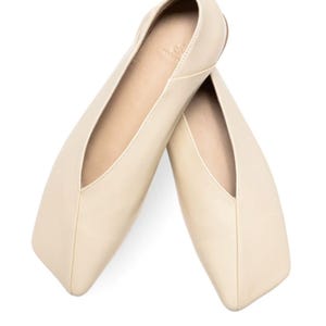 May include: A pair of cream-colored ballet flats with a square toe and a low-cut vamp. The shoes are made of a smooth leather-like material and are set against a white background.