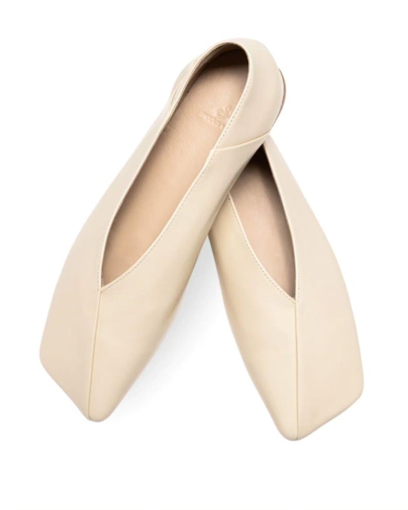 May include: A pair of cream-colored ballet flats with a square toe and a low-cut vamp. The shoes are made of a smooth leather-like material and are set against a white background.