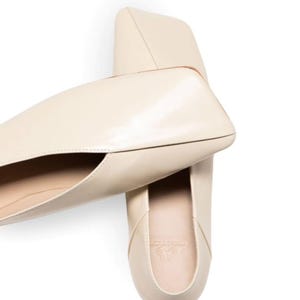 May include: Cream-colored leather flats with a unique, angular design. The shoes have a pointed toe and a low heel. The top-down view showcases the shoe's sleek silhouette and minimalist aesthetic. The flats are likely designed for comfort and style.