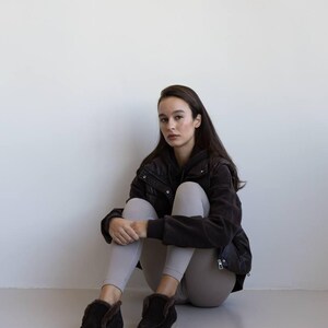 Op de afbeelding: Een persoon draagt een zwarte jas, grijze leggings en zwarte su&egrave;de laarzen met imitatiebont. De laarzen hebben een laag profiel en een neutrale zool. De persoon zit op een lichtgrijze ondergrond.