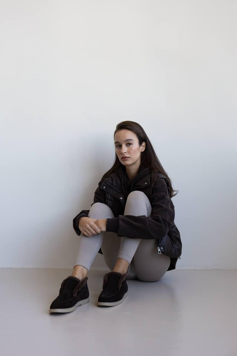 Op de afbeelding: Een persoon draagt een zwarte jas, grijze leggings en zwarte su&egrave;de laarzen met imitatiebont. De laarzen hebben een laag profiel en een neutrale zool. De persoon zit op een lichtgrijze ondergrond.
