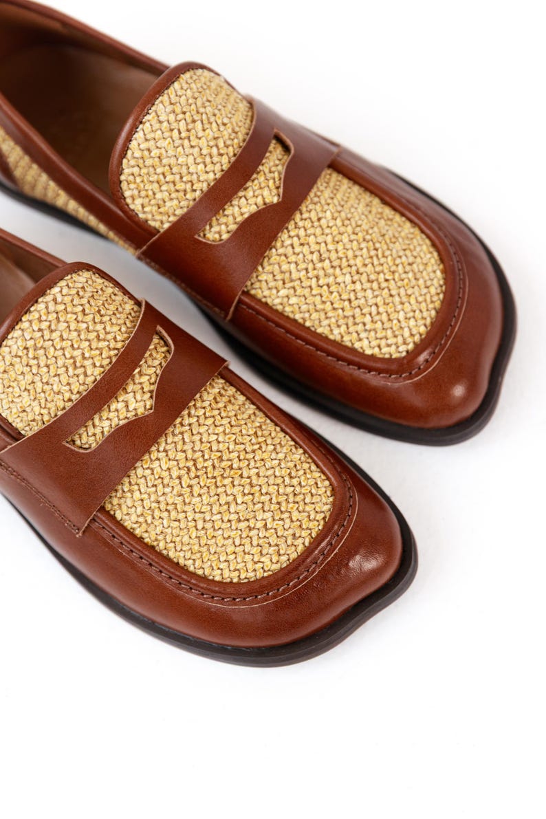 May include: A pair of brown leather loafers with woven straw-colored panels. The shoes have a classic penny loafer design with a rounded toe and a dark brown sole. The shoes are displayed on a white background.