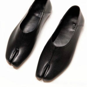 Könnte beinhalten: Ein Paar schwarze Leder-Split-Toe-Flats. Die Schuhe haben ein minimalistisches Design mit einer Trennung zwischen dem großen Zeh und den anderen Zehen. Die Flats befinden sich auf weißem Hintergrund.