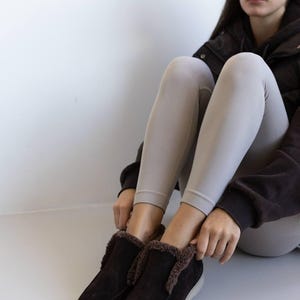 Op de afbeelding: Donkerbruine su&egrave;de instappers met een imitatiebontvoering en een witte zool. De schoenen zijn ontworpen voor comfort en warmte. Het schoeisel is geschikt voor binnen of buiten.
