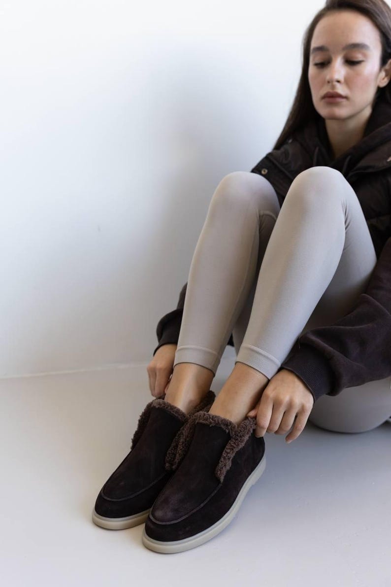 Op de afbeelding: Donkerbruine su&egrave;de instappers met een imitatiebontvoering en een witte zool. De schoenen zijn ontworpen voor comfort en warmte. Het schoeisel is geschikt voor binnen of buiten.