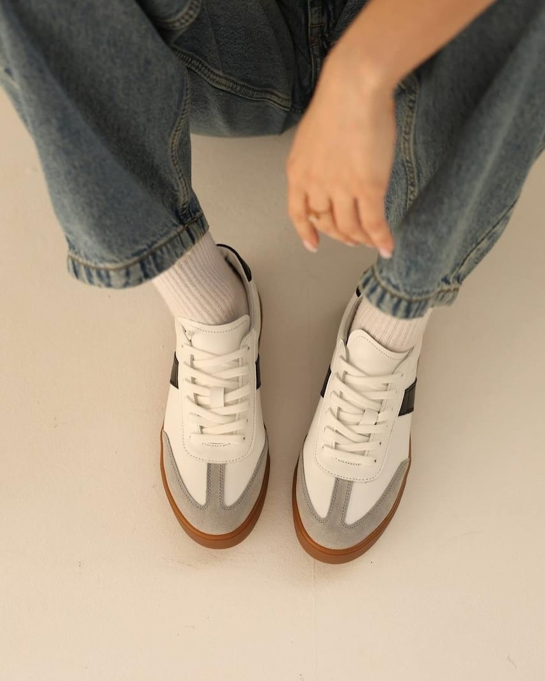 May include: White leather sneakers with black and gray accents and brown soles. The sneakers have a classic design with laces and a rounded toe.