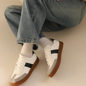 May include: White leather sneakers with black stripes and a gum sole. The sneakers are worn with white socks and blue jeans.