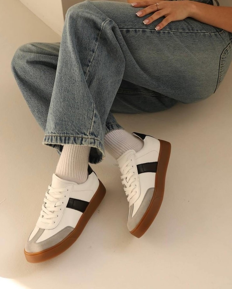 May include: White leather sneakers with black stripes and a gum sole. The sneakers are worn with white socks and blue jeans.