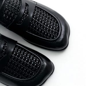 May include: Pair of black leather loafers with woven detailing on the vamp and strap. The shoes have a classic design with a rounded toe and a low heel. The loafers are set against a white background.
