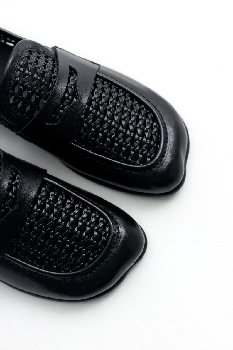 May include: Pair of black leather loafers with woven detailing on the vamp and strap. The shoes have a classic design with a rounded toe and a low heel. The loafers are set against a white background.