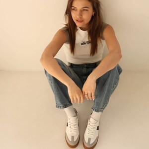 May include: A person wearing a white and gray tank top with the text "Two Seas New York" printed on the front, blue jeans, and white sneakers with black accents.