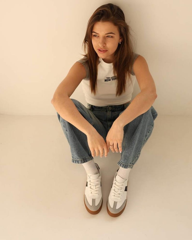 May include: A person wearing a white and gray tank top with the text "Two Seas New York" printed on the front, blue jeans, and white sneakers with black accents.