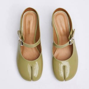 May include: A pair of olive green, patent leather Mary Jane style shoes. Each shoe has a Tabi toe, a strap across the top with a silver buckle, and a small heel. The interior is a light brown color.