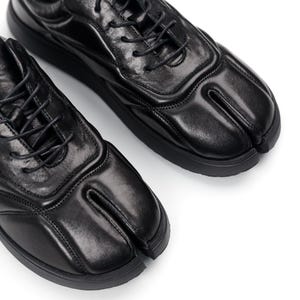 Op de afbeelding: Paar zwarte leren schoenen met split-teen en veters. De schoenen hebben een uniek ontwerp met een apart gedeelte voor de grote teen. De zolen zijn zwart en de schoenen staan op een witte achtergrond.