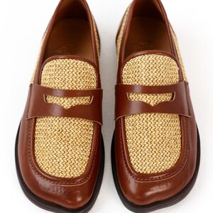 May include: A pair of brown leather loafers with woven straw-colored panels. The shoes have a classic penny loafer design with a strap across the vamp. The soles are dark brown.