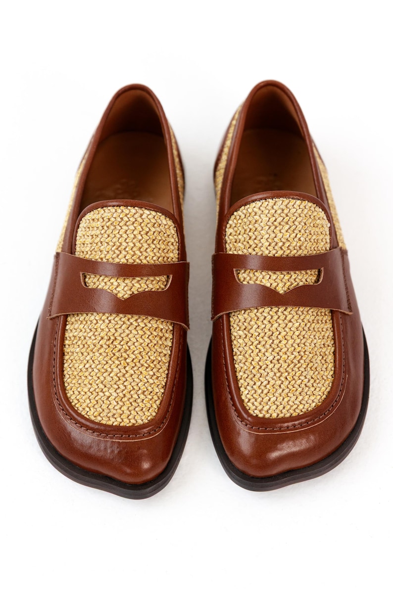 May include: A pair of brown leather loafers with woven straw-colored panels. The shoes have a classic penny loafer design with a strap across the vamp. The soles are dark brown.