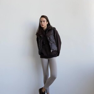 Op de afbeelding: Een persoon draagt een zwarte gewatteerde bodywarmer over een donkerbruine hoodie, gecombineerd met lichtgrijze leggings en zwarte schoenen. De outfit is casual en eenvoudig, geschikt voor dagelijks gebruik. De achtergrond is een witte muur.