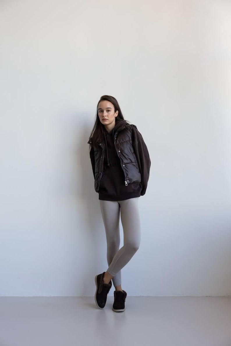 Op de afbeelding: Een persoon draagt een zwarte gewatteerde bodywarmer over een donkerbruine hoodie, gecombineerd met lichtgrijze leggings en zwarte schoenen. De outfit is casual en eenvoudig, geschikt voor dagelijks gebruik. De achtergrond is een witte muur.