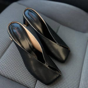 K&ouml;nnte beinhalten: Ein Paar schwarze Leder-Mules mit eckiger Zehenpartie und kleinem Blockabsatz. Die Innenseite ist hellbraun und der obere Rand ist goldfarben eingefasst. Der Markenname "Amatto" ist auf der Innensohle aufgedruckt.