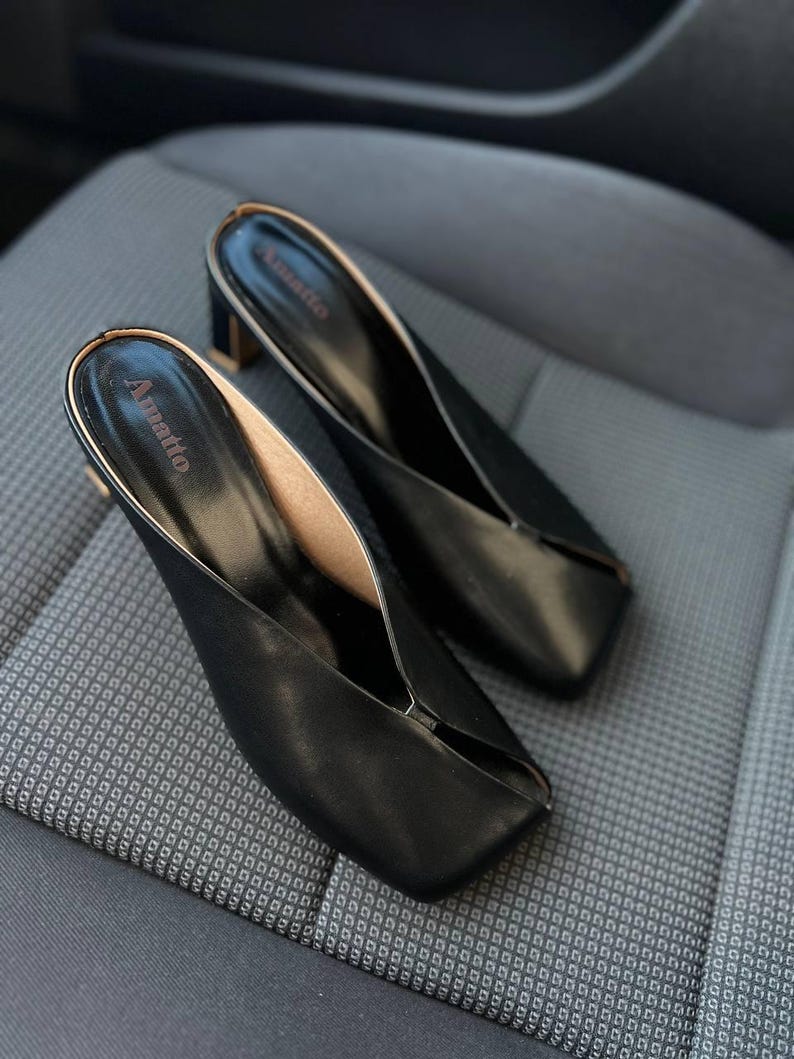 K&ouml;nnte beinhalten: Ein Paar schwarze Leder-Mules mit eckiger Zehenpartie und kleinem Blockabsatz. Die Innenseite ist hellbraun und der obere Rand ist goldfarben eingefasst. Der Markenname "Amatto" ist auf der Innensohle aufgedruckt.