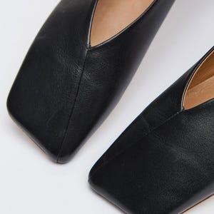 May include: Close-up of two black leather flats with a square toe design. The shoes have a low-cut vamp and a smooth, matte finish. The image highlights the sleek, minimalist style of the footwear.