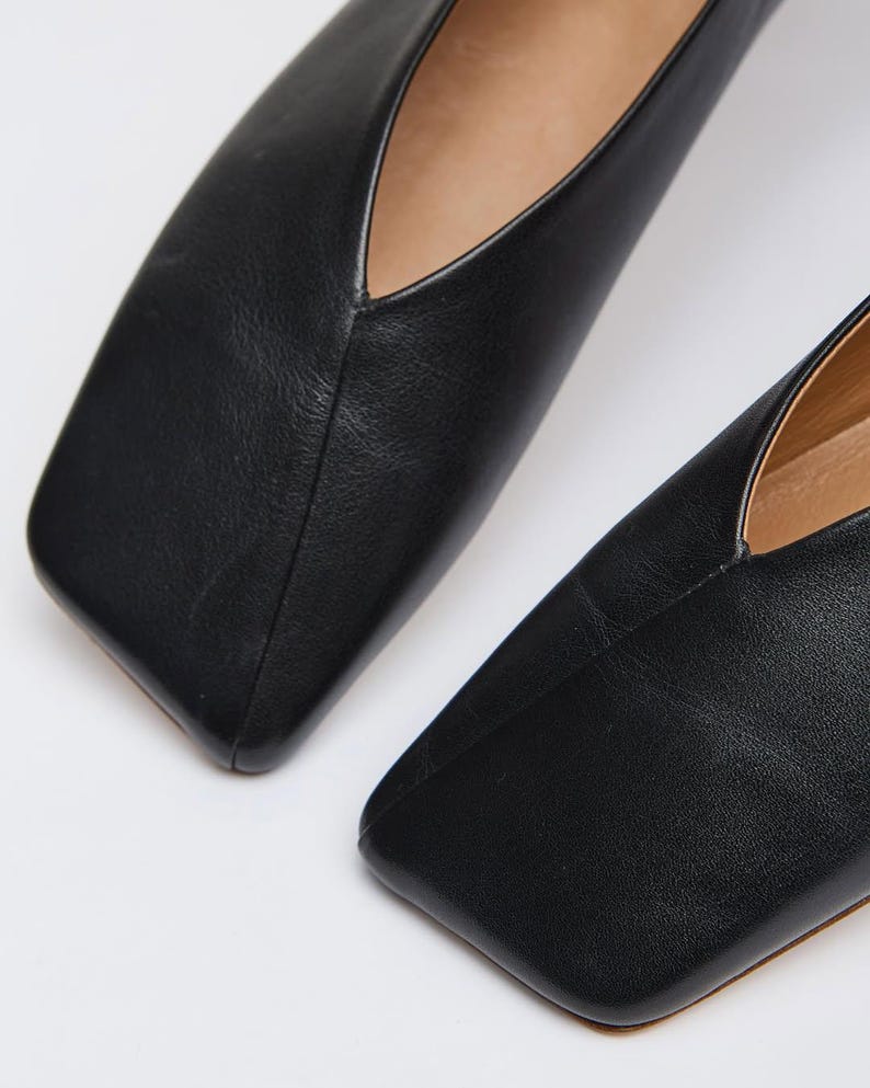 May include: Close-up of two black leather flats with a square toe design. The shoes have a low-cut vamp and a smooth, matte finish. The image highlights the sleek, minimalist style of the footwear.