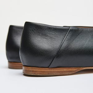May include: Close-up of two black leather flats with a low heel and a wooden sole. The shoes have a simple, classic design, with a smooth, matte finish. The heels are approximately 0.5 inches high.