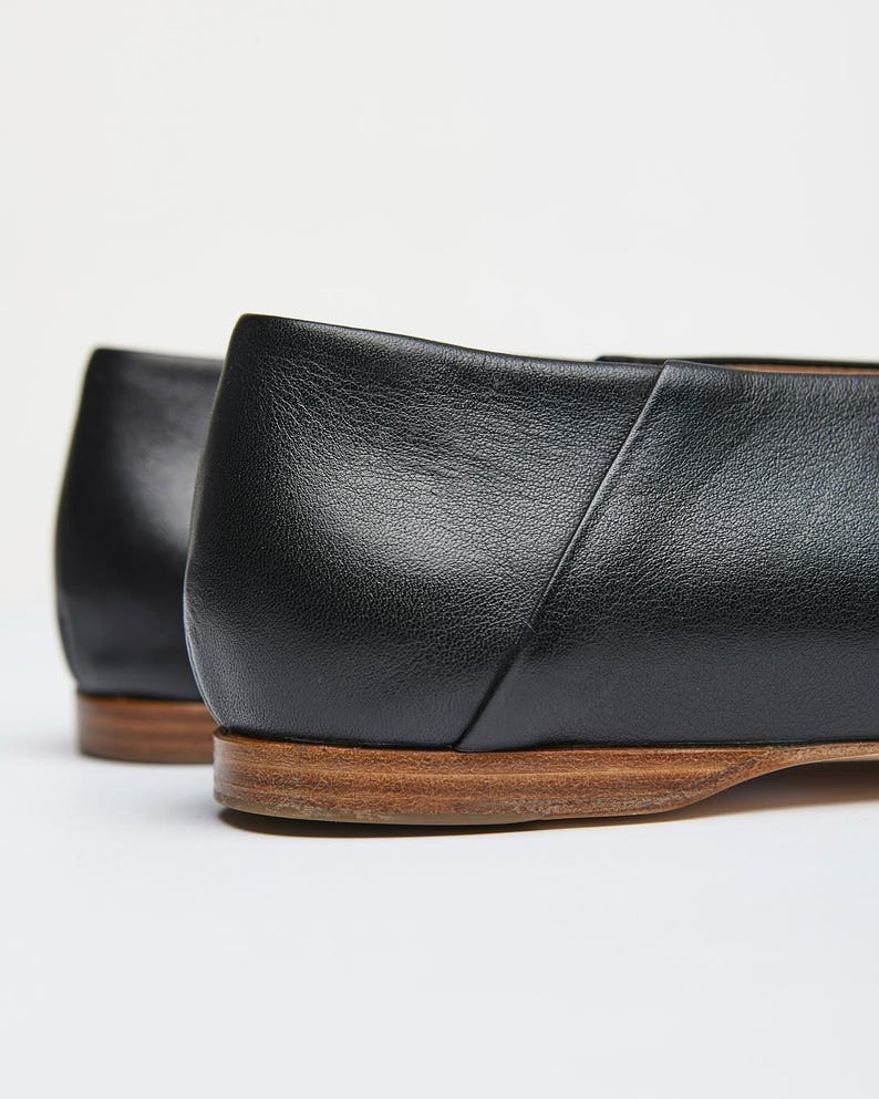 May include: Close-up of two black leather flats with a low heel and a wooden sole. The shoes have a simple, classic design, with a smooth, matte finish. The heels are approximately 0.5 inches high.