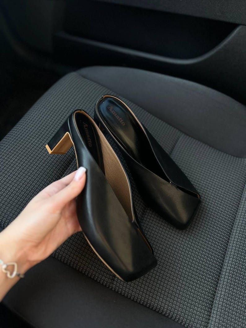 K&ouml;nnte beinhalten: Schwarze Leder-Mules mit eckigen Zehen und Blockabs&auml;tzen. Die Schuhe haben ein minimalistisches Design, mit dem Markennamen "Amatto" auf der Innensohle. Die Abs&auml;tze haben einen goldfarbenen Akzent.