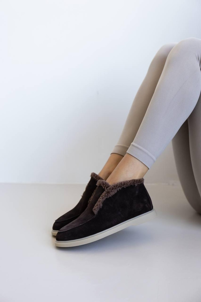 Op de afbeelding: Donkerbruine su&egrave;de enkellaarsjes met een cr&egrave;mekleurige zool en imitatiebontvoering. De laarzen worden getoond met lichtgrijze leggings. Deze comfortabele laarzen zijn ideaal voor dagelijks gebruik.
