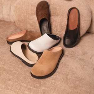 May include: An assortment of leather and suede slip-on shoes in various neutral colors, including black, white, brown, and beige. The shoes are arranged on a textured, light brown surface, showcasing their different styles and materials.
