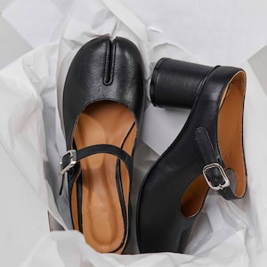 May include: A pair of black leather heeled shoes with a unique toe design. The shoes have a buckle strap and a chunky heel.
