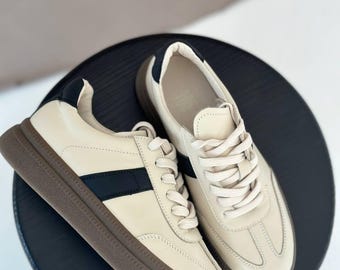 May include: A pair of cream-colored sneakers with a black stripe and brown soles. The shoes have a classic design with laces and a padded collar. The sneakers are displayed on a black surface.
