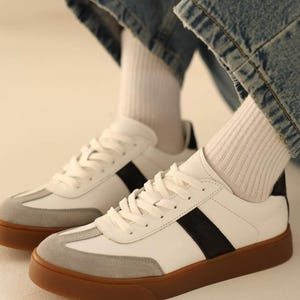 May include: White leather sneakers with black stripes and a gum sole. The sneakers have a suede toe cap and are worn with white socks and blue jeans.