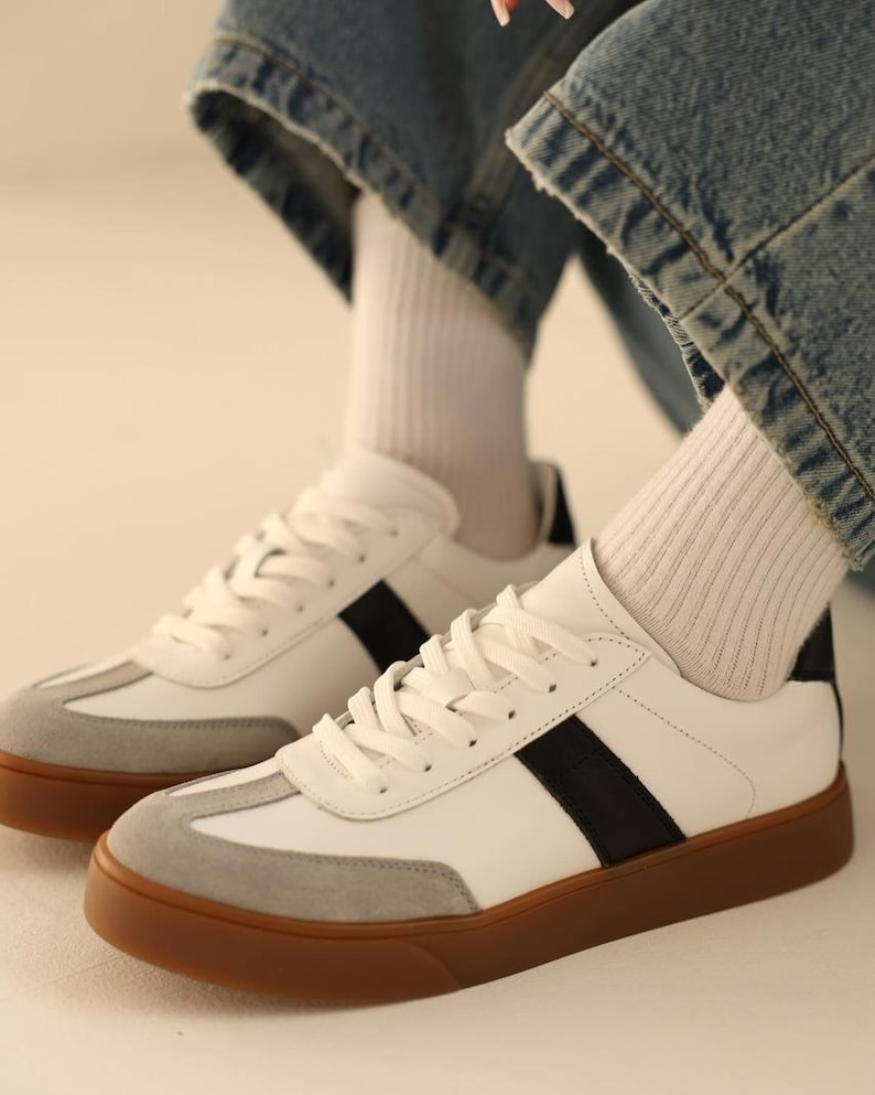May include: White leather sneakers with black stripes and a gum sole. The sneakers have a suede toe cap and are worn with white socks and blue jeans.