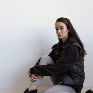 Op de afbeelding: Een persoon draagt een zwarte gewatteerde bodywarmer en bijpassende zwarte schoenen met een voering van imitatiebont. De persoon draagt ook lichtgrijze leggings en een zwarte hoodie. De achtergrond is een effen witte muur.