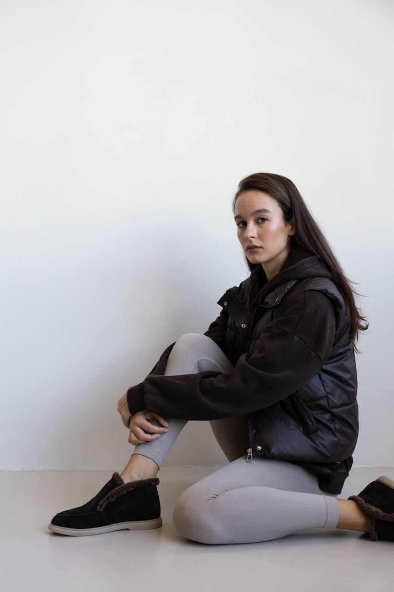 Op de afbeelding: Een persoon draagt een zwarte gewatteerde bodywarmer en bijpassende zwarte schoenen met een voering van imitatiebont. De persoon draagt ook lichtgrijze leggings en een zwarte hoodie. De achtergrond is een effen witte muur.