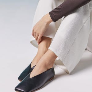 May include: Black leather flats with a square toe and a V-shaped vamp. The shoes have a low heel and a minimalist design. The flats are paired with white textured pants and a brown long-sleeve top.