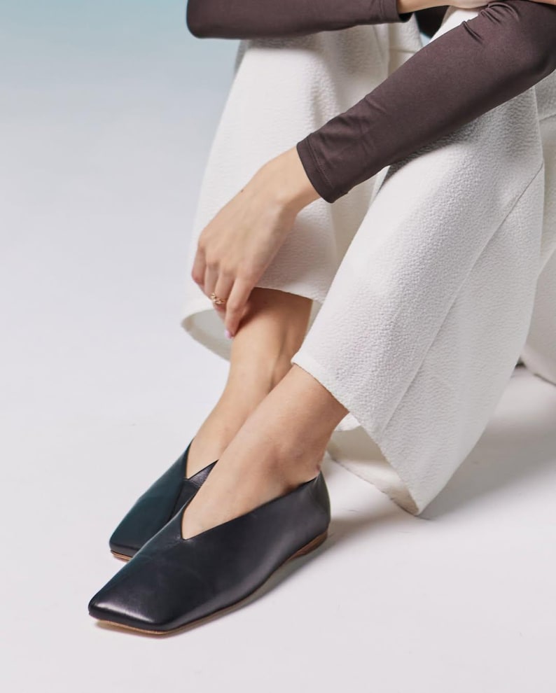 May include: Black leather flats with a square toe and a V-shaped vamp. The shoes have a low heel and a minimalist design. The flats are paired with white textured pants and a brown long-sleeve top.