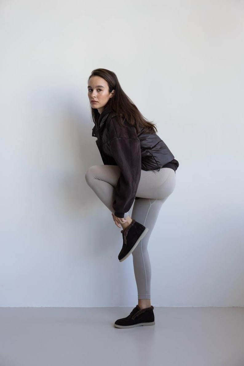 Op de afbeelding: Een persoon draagt een zwarte pufferjack over een donkerbruine fleecejas, lichtgrijze leggings en zwarte su&egrave;de enkellaarsjes met een voering van imitatiebont. De persoon buigt zich en raakt de enkellaars aan.
