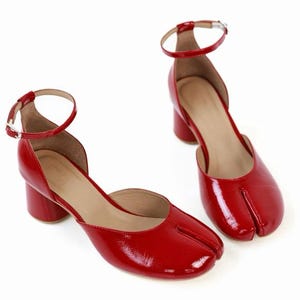 May include: Pair of red patent leather heeled shoes with a unique split-toe design. The shoes have a low block heel and an ankle strap with a silver buckle. The shoes are a vibrant red color.