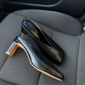 K&ouml;nnte beinhalten: Schwarze Leder-Mules mit eckiger Zehenpartie und kleinem, goldfarbenem Blockabsatz. Die Schuhe sind auf einer gemusterten, grauen Oberfl&auml;che platziert. Der Markenname "Anatto" ist im Inneren eines Schuhs sichtbar. Die Abs&auml;tze sind ca. 5 cm hoch.