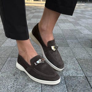 May include: Brown suede loafers with a white sole. Each shoe features a decorative silver buckle. The shoes are a slip-on style and appear to be made of a soft suede material. The shoes are shown on a gray tiled surface.